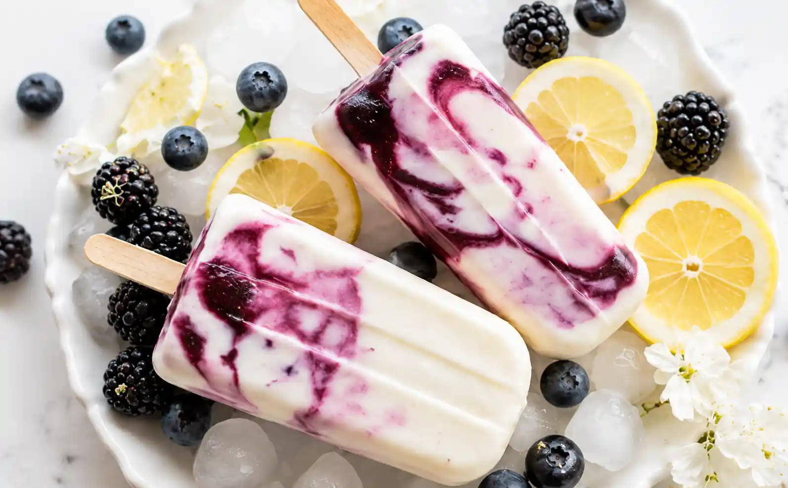 Creamy berry lemon yogurt popsicles with blackberry swirls, blueberries, lemon slices, ice cubes, and white blossoms on a plate